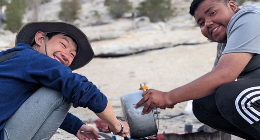 two students smile at the camera as they prepare food on a backpacking trip